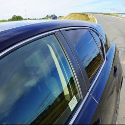 BLACK STUDIO - Ciemny samochód z naklejką na szybie, odbicie nieba w szybach. Widok z boku, asfaltowa droga w tle.