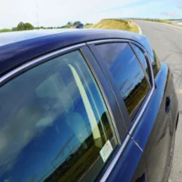 Ciemny samochód z naklejką na szybie, odbicie nieba w szybach. Widok z boku, asfaltowa droga w tle.