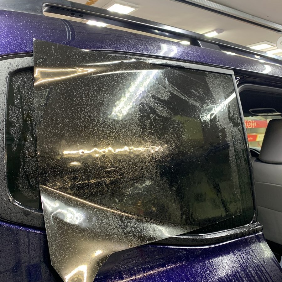 Proces przyciemniania tylnej szyby samochodu folią, widoczne drobne pęcherzyki powietrza i woda, niebieski lakier karoserii.