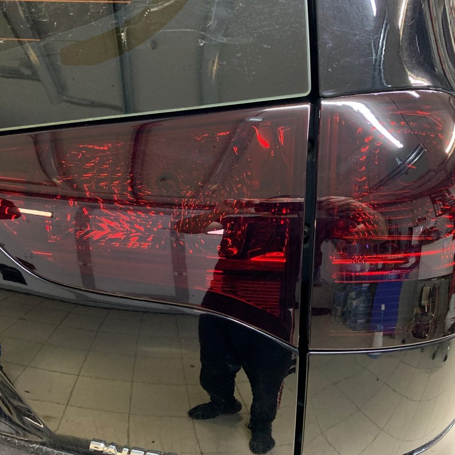 Ciemnoczerwone, przyciemnione tylne światło czarnego samochodu. Widoczne odbicie osoby wykonującej usługę.