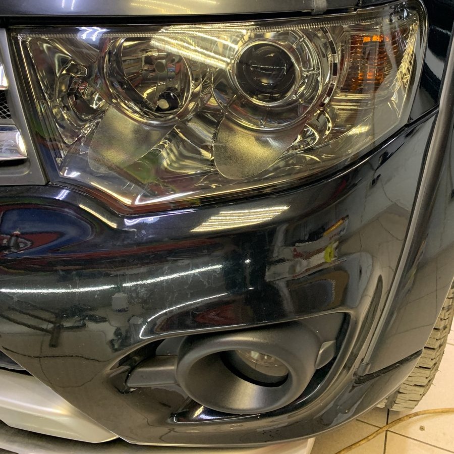 Błyszczący reflektor i fragment karoserii czarnego samochodu przed zabiegiem renowacji, widoczne drobne zarysowania i zabrudzenia na lakierze.