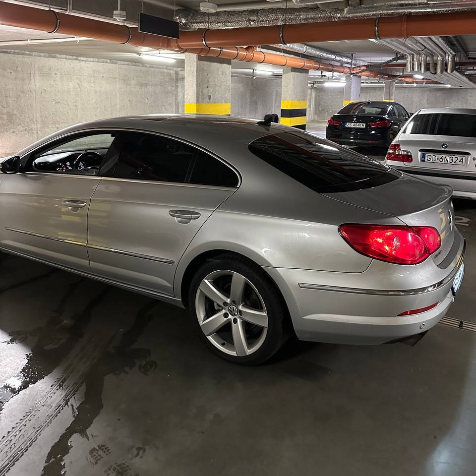 Srebrny Volkswagen Passat CC z przyciemnionymi szybami zaparkowany w garażu podziemnym, widok z tyłu i boku, oświetlenie sufitowe odbija się w lakierze.