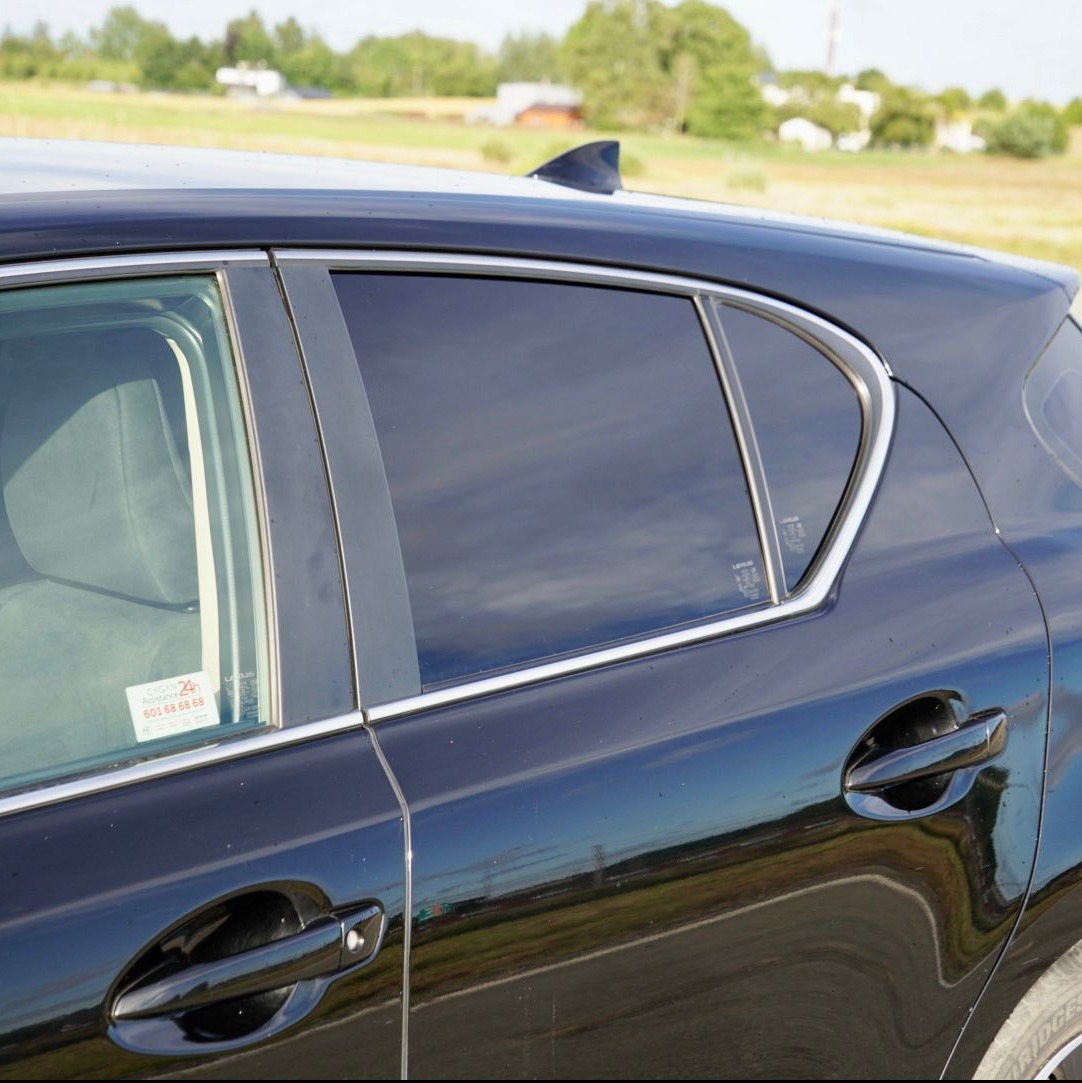 Ciemne szyby w czarnym samochodzie marki Lexus, widoczne z boku na tle zieleni. Przyciemnione tylne szyby, chromowane obramowania okien, antena rekinka.