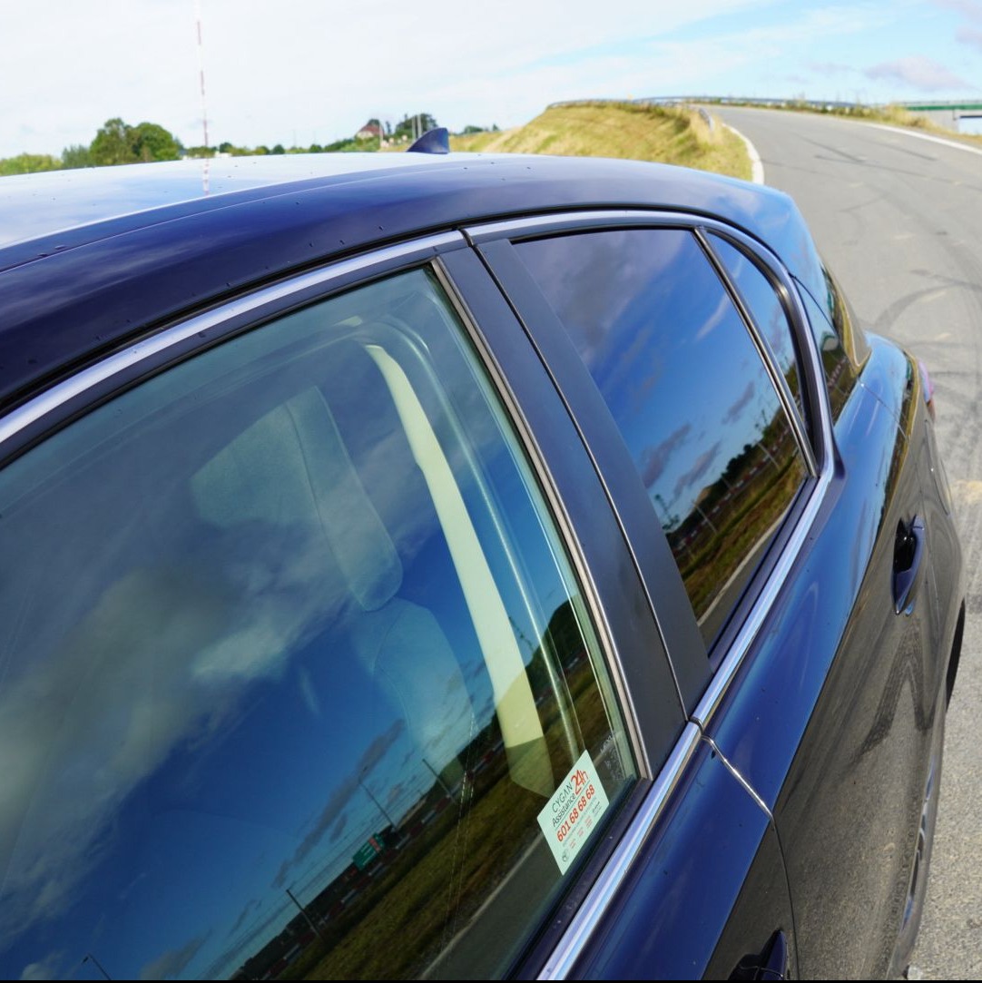 Ciemny samochód z naklejką na szybie, odbicie nieba w szybach. Widok z boku, asfaltowa droga w tle.