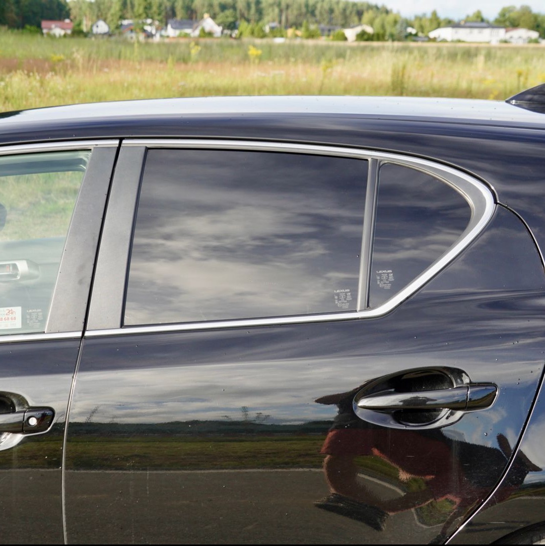 Ciemne szyby w czarnym samochodzie Lexus, odbijające chmury. Widoczne naklejki na szybie. Tło to zielona łąka i drzewa.