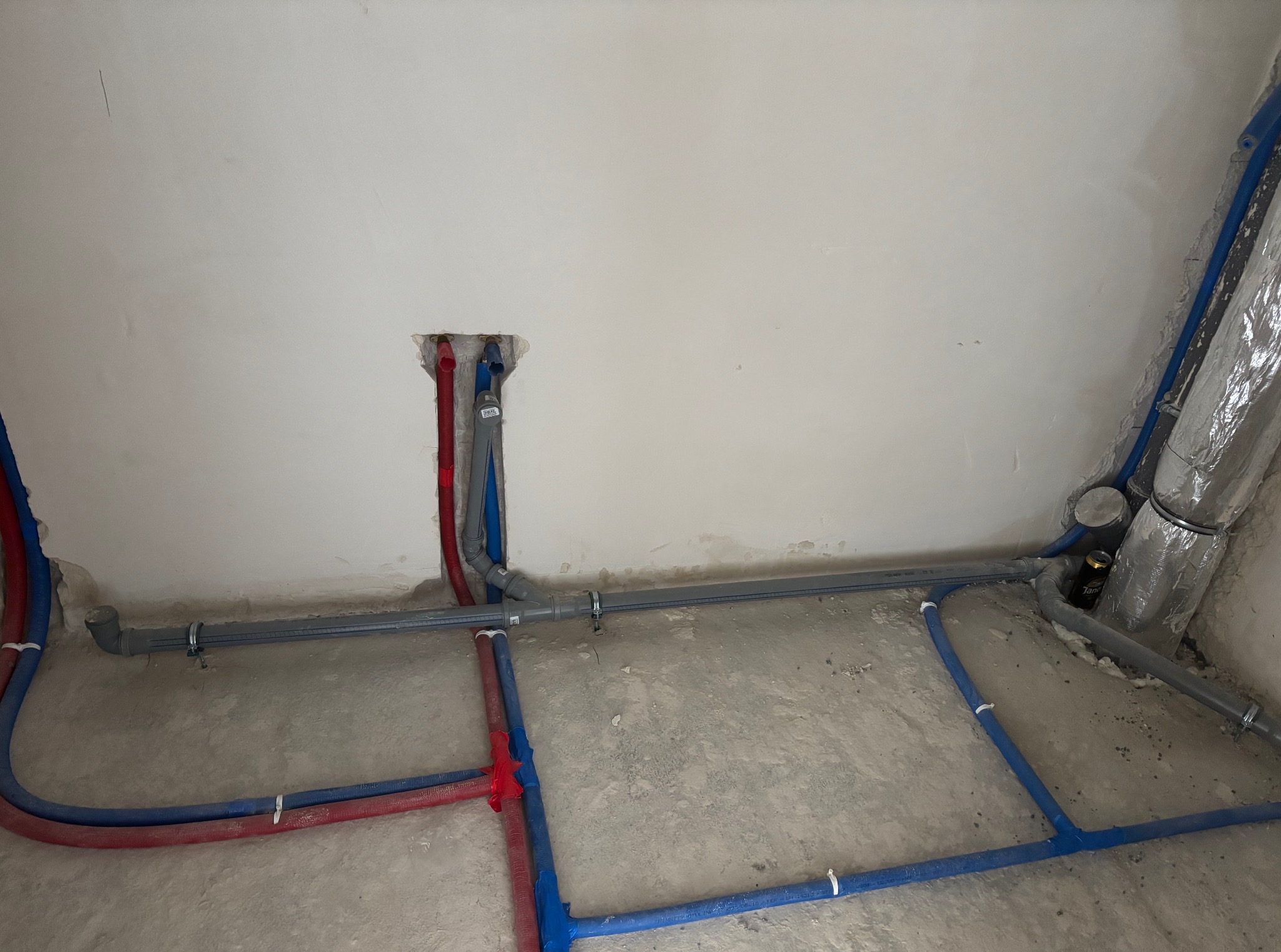 Instalacja wodna i kanalizacyjna w surowym stanie: rury czerwone i niebieskie przy ścianie, szare rury odpływowe pod sufitem i na podłodze, przed wylaniem posadzki.