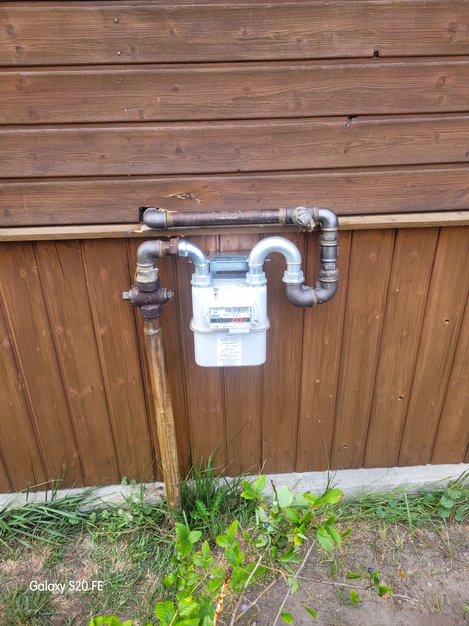 Zamontowany gazomierz na brązowej, drewnianej ścianie budynku. Widoczne rury doprowadzające gaz. U dołu roślinność.