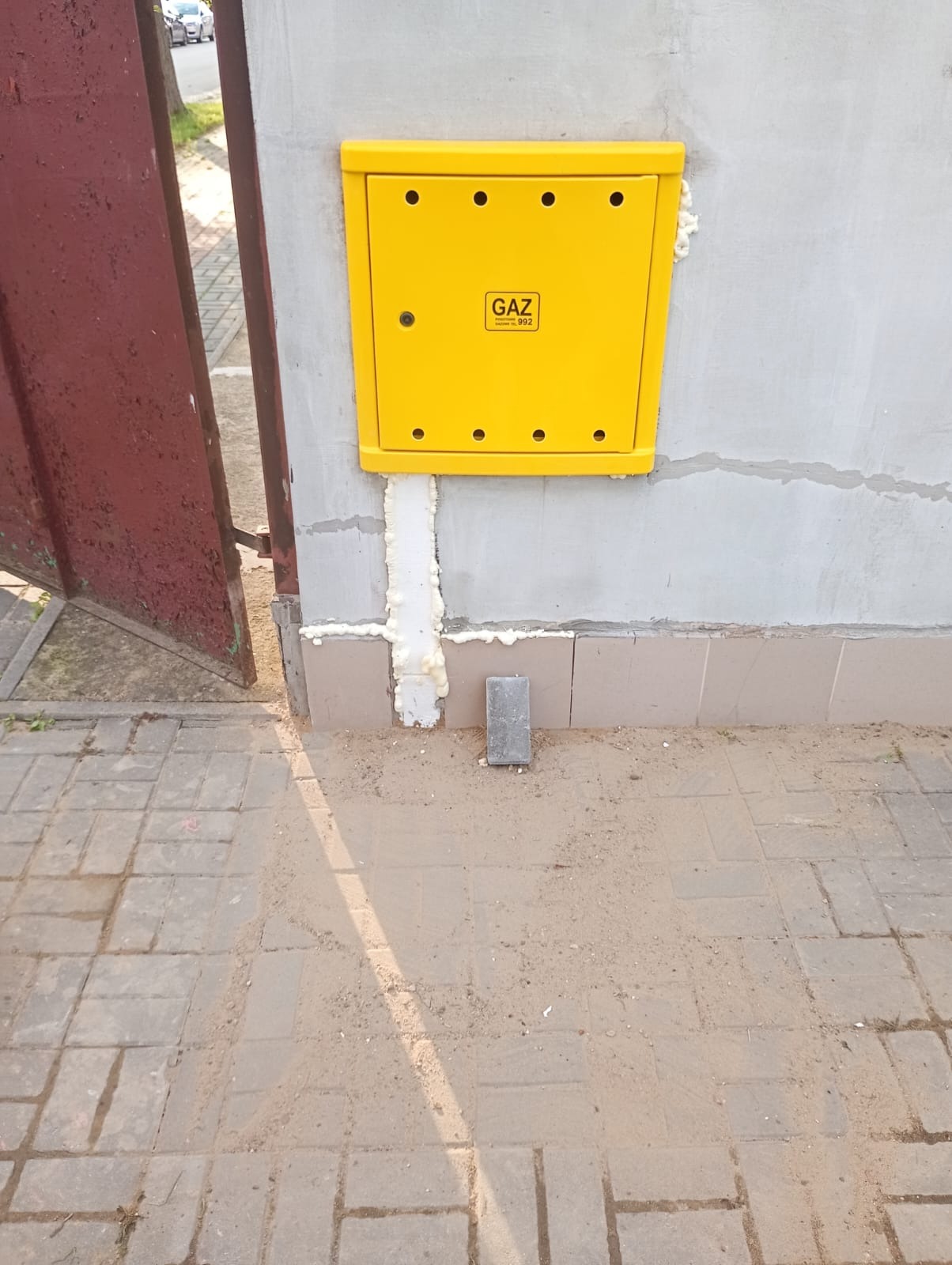 Żółta skrzynka gazowa na szarej ścianie budynku, zabezpieczona pianką montażową. Obok fragment czerwonej bramy i kostka brukowa.