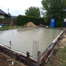 MB USŁUGI W BUDOWNICTWIE BRONIOWSKI MACIEJ - Zalany fundament pod budowę domu, z widocznym zbrojeniem i szalunkiem. W tle zielony trawnik, drzewa i przenośna toaleta. Widoczny niwelator laserowy.