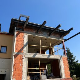 MB USŁUGI W BUDOWNICTWIE BRONIOWSKI MACIEJ - Budowa domu z czerwonej cegły, widoczne stalowe belki podtrzymujące strop i rusztowanie. Częściowo otynkowana fasada i błękitne niebo w tle.