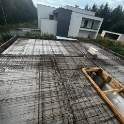 MB USŁUGI W BUDOWNICTWIE BRONIOWSKI MACIEJ - Zbrojenie stropu budynku jednorodzinnego w Jaworznie, przygotowane do zalania betonem. Widoczne stalowe pręty i szalunki okienne. W tle nowoczesny dom.