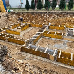 MB USŁUGI W BUDOWNICTWIE BRONIOWSKI MACIEJ - Zbrojenie i szalunek fundamentów domu jednorodzinnego, widok z góry. Na drugim planie pracownik przy pracy, ziemia, budynek i zieleń.