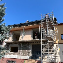 MB USŁUGI W BUDOWNICTWIE BRONIOWSKI MACIEJ - Budowa domu wielorodzinnego w trakcie realizacji. Widoczne rusztowania, pracownicy na dachu i elewacja w stanie surowym z oknami bez szyb. Duże drzewo z lewej strony.