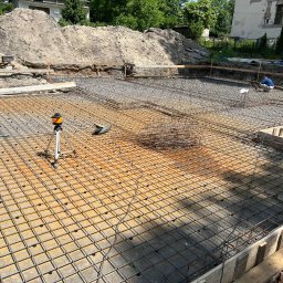 MB USŁUGI W BUDOWNICTWIE BRONIOWSKI MACIEJ - Zbrojenie fundamentów domu jednorodzinnego. Widoczna siatka z prętów, szalunki i niwelator laserowy. W tle hałda ziemi.