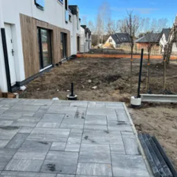 Nowoczesny dom w zabudowie szeregowej z wybrukowanym tarasem i przygotowanym terenem pod ogród. Widoczne młode drzewka i elementy konstrukcyjne.