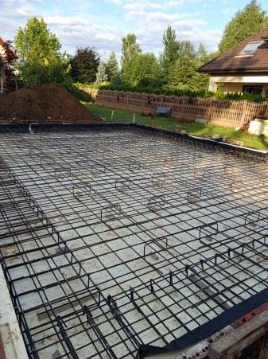 Zbrojenie płyty fundamentowej na przygotowanym podłożu, widoczne elementy szalunku i otoczenie działki z zielenią. Ujęcie z góry.