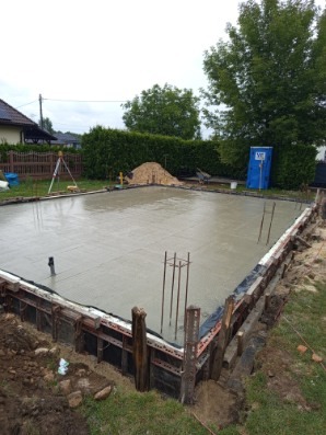 Zalany fundament pod budowę domu, z widocznym zbrojeniem i szalunkiem. W tle zielony trawnik, drzewa i przenośna toaleta. Widoczny niwelator laserowy.