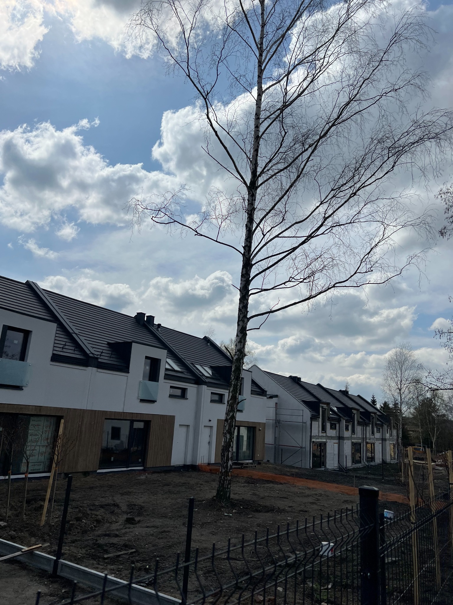 Szereg domów jednorodzinnych z ciemnymi dachami i jasnymi elewacjami, widziane zza ogrodzenia. W tle pochmurne niebo i drzewo bez liści.