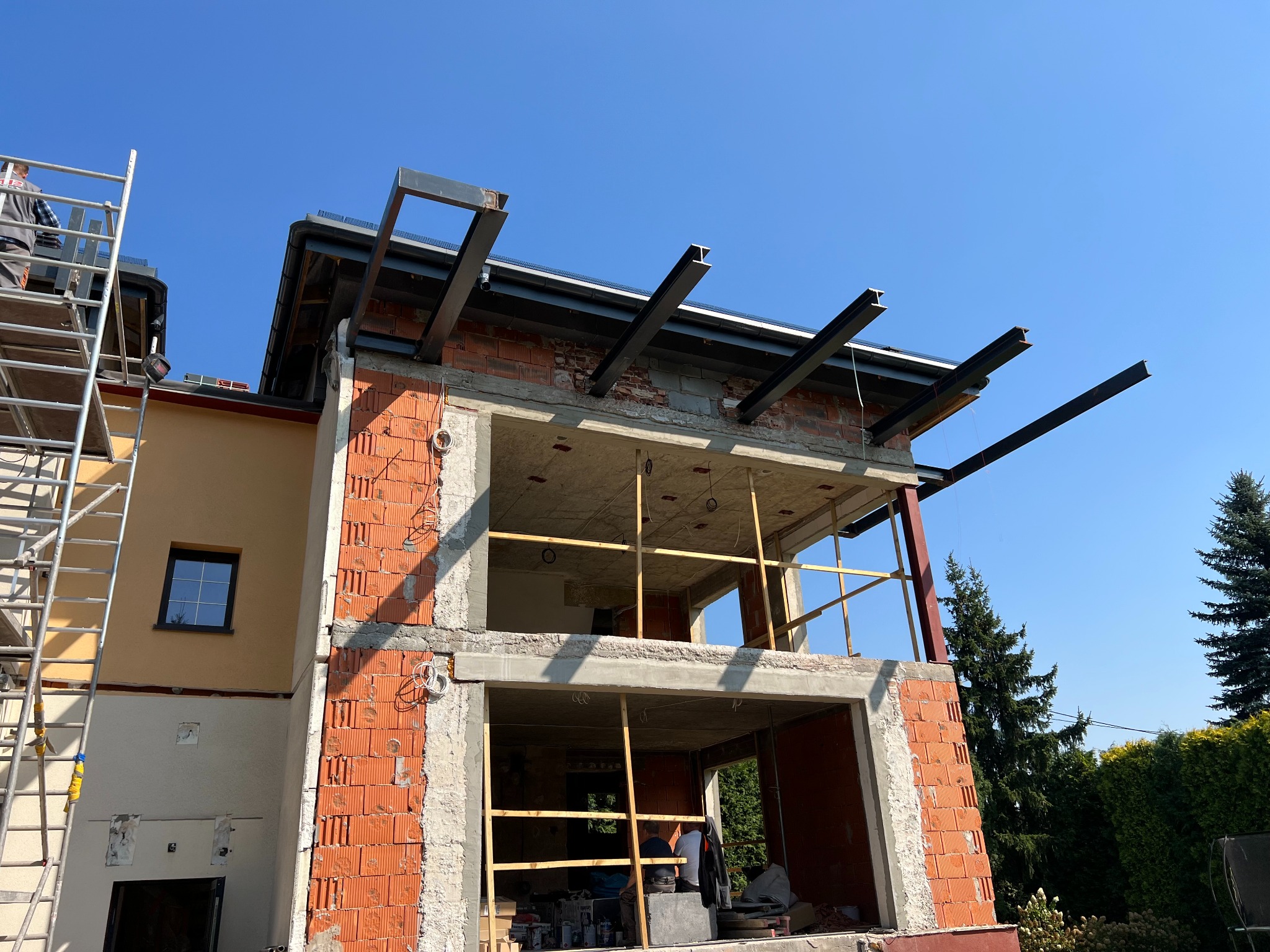 Budowa domu z czerwonej cegły, widoczne stalowe belki podtrzymujące strop i rusztowanie. Częściowo otynkowana fasada i błękitne niebo w tle.