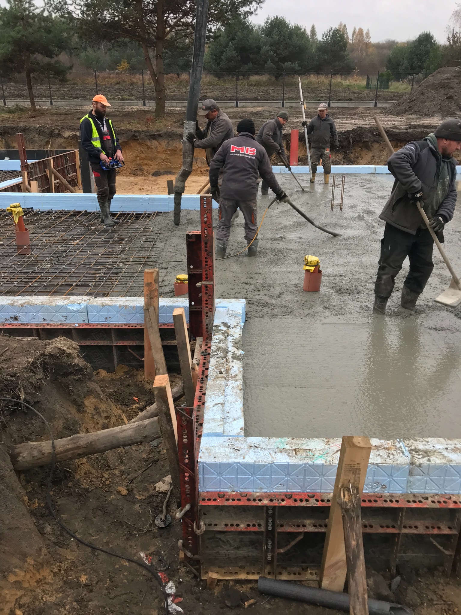 Wylewanie betonu na fundamenty z użyciem pompy. Pracownicy w kaloszach rozprowadzają beton. Widoczne zbrojenie, szalunki i wykop pod fundament. Praca w toku.