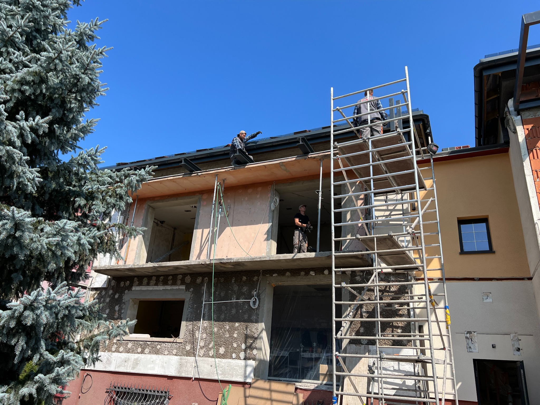 Budowa domu wielorodzinnego w trakcie realizacji. Widoczne rusztowania, pracownicy na dachu i elewacja w stanie surowym z oknami bez szyb. Duże drzewo z lewej strony.