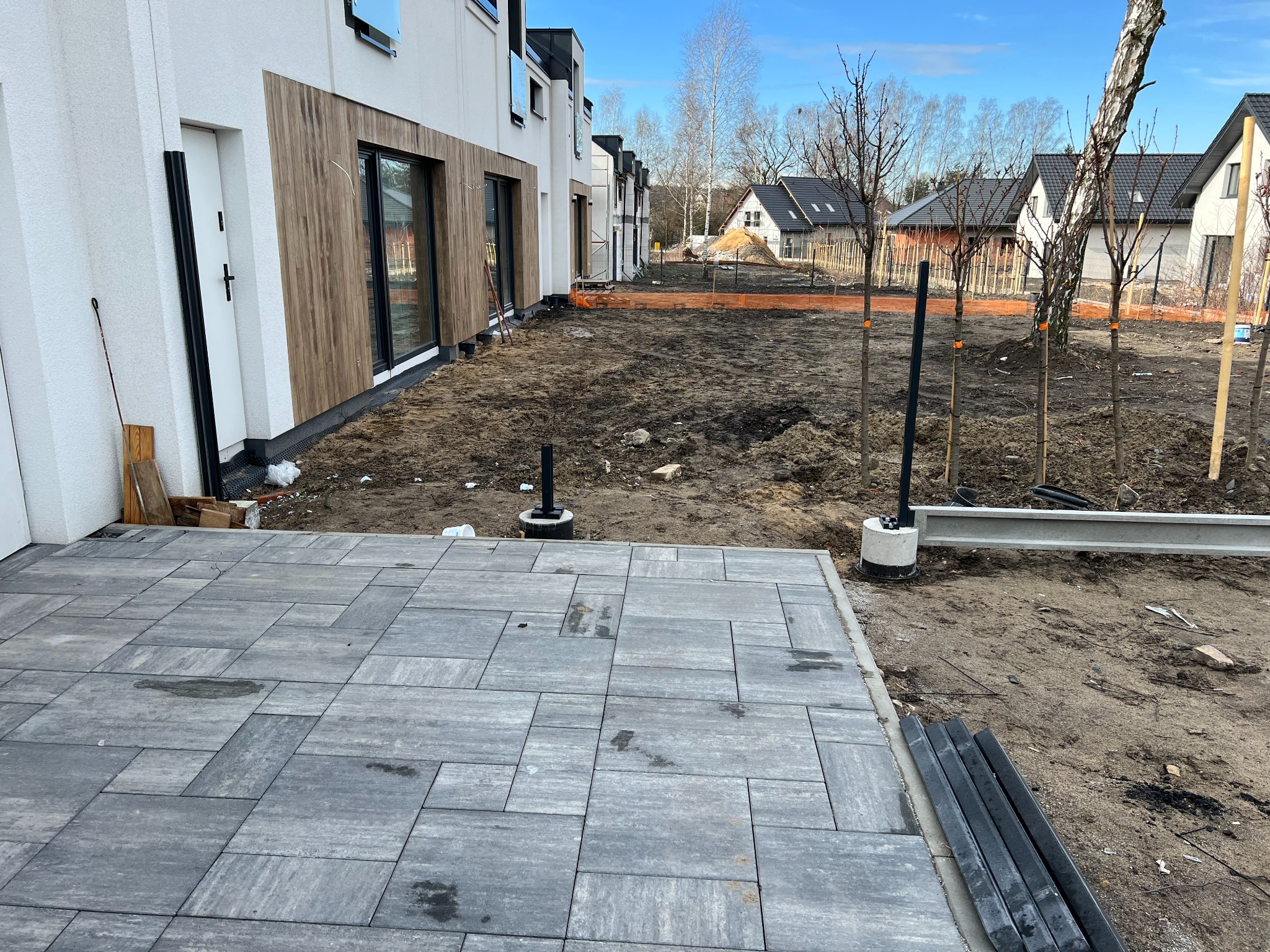 Nowoczesny dom w zabudowie szeregowej z wybrukowanym tarasem i przygotowanym terenem pod ogród. Widoczne młode drzewka i elementy konstrukcyjne.