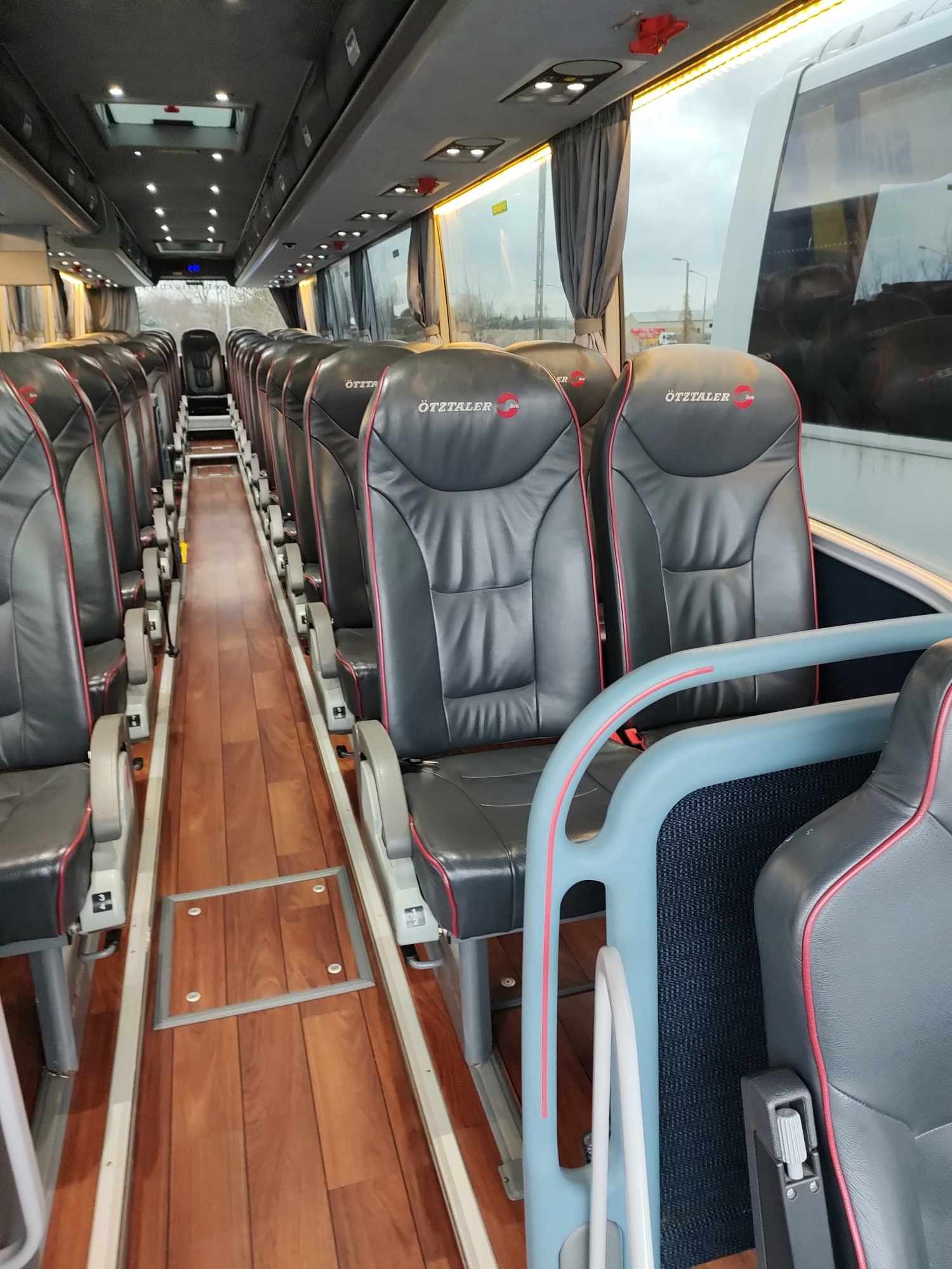 Wnętrze luksusowego autobusu z czarnymi skórzanymi fotelami z czerwonymi akcentami i logo Ötztaler, drewnianą podłogą i dużymi oknami.