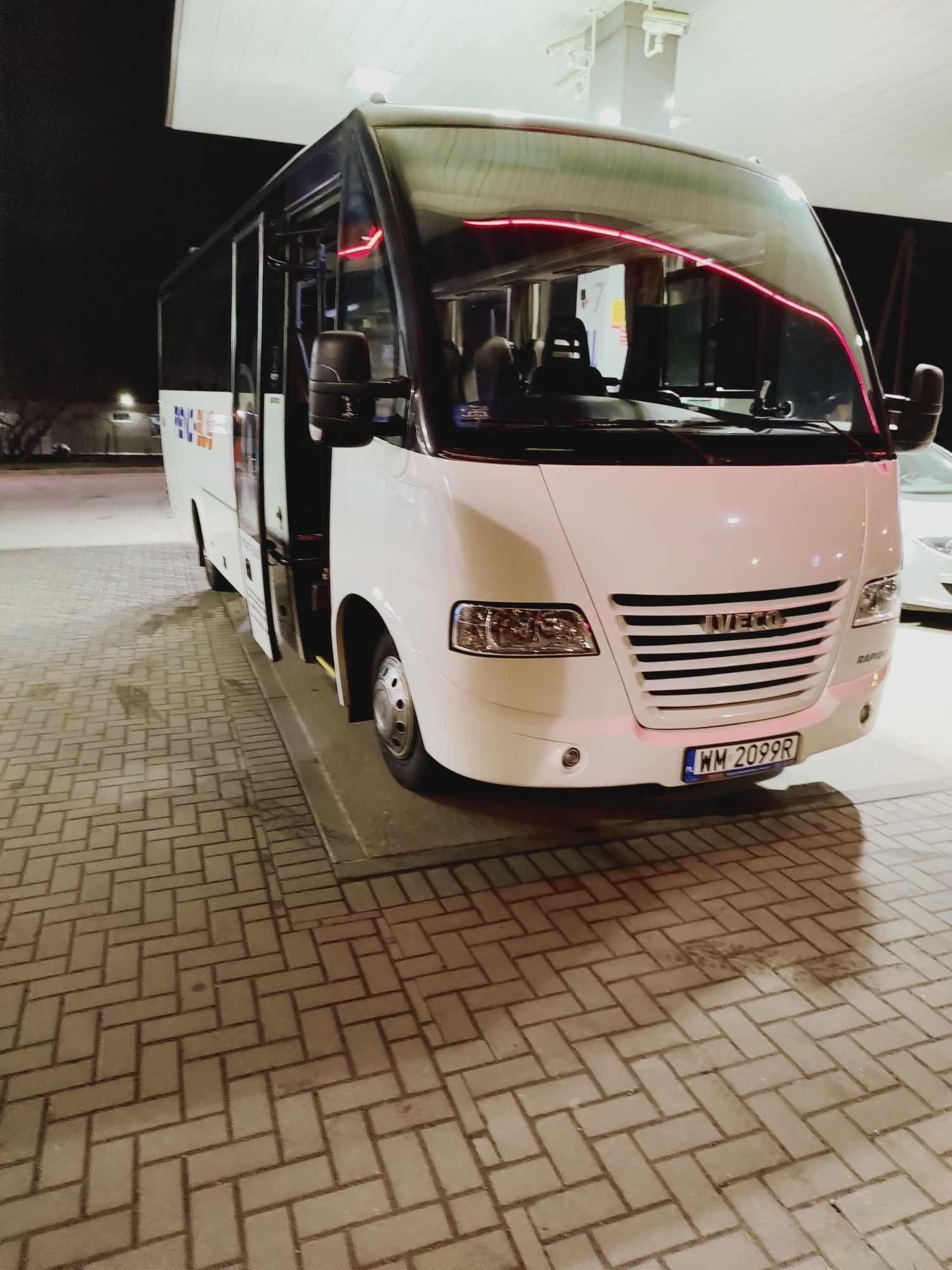 Biały bus Iveco z otwartymi drzwiami, zaparkowany na kostce brukowej. Widoczna tablica rejestracyjna WM 2099R i neonowe oświetlenie wnętrza. Nocne ujęcie z przodu.