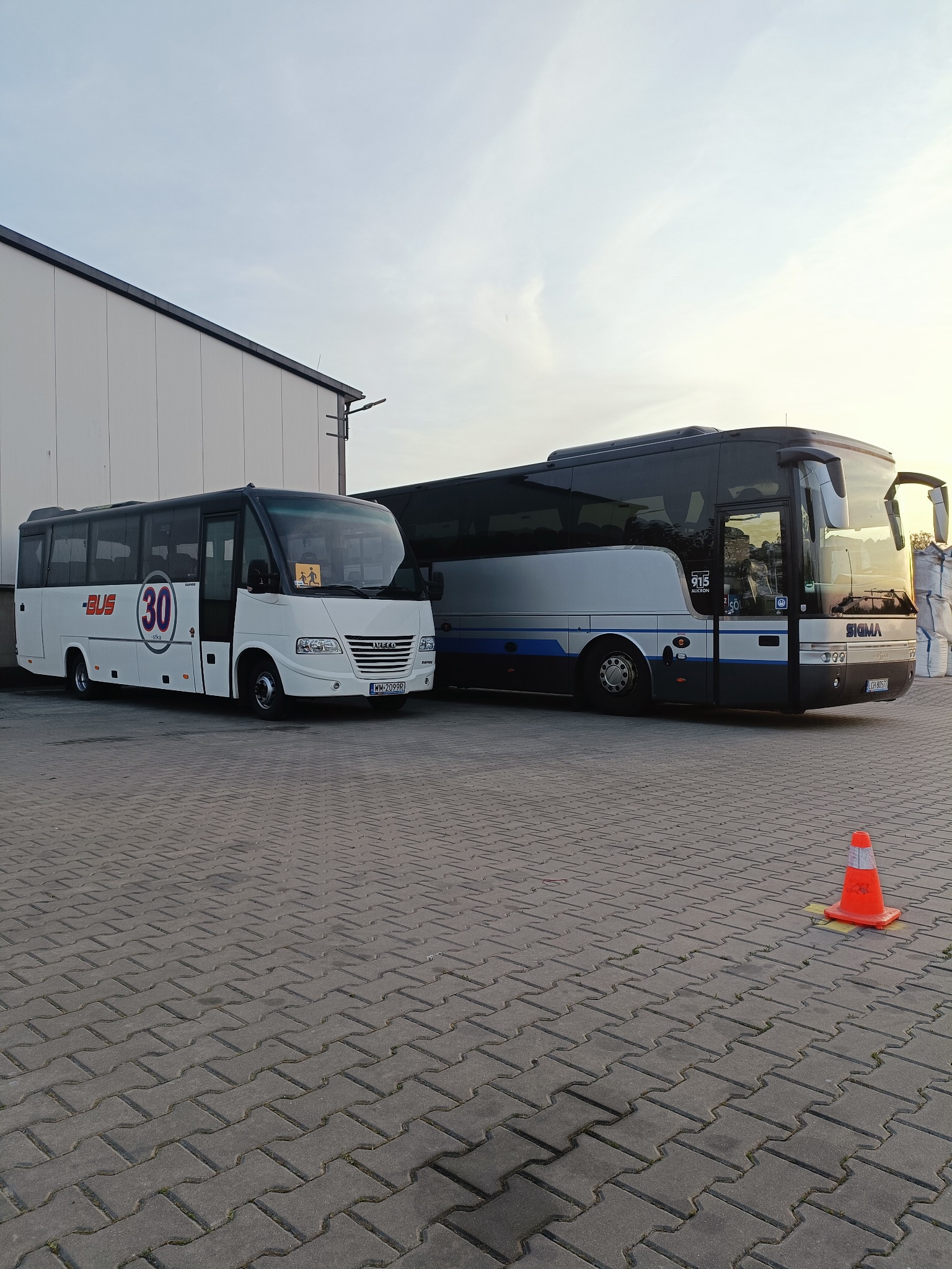 Dwa autobusy: biały z numerem 30 i ciemny z napisem SIGMA, stojące na placu z kostki, z pomarańczowym pachołkiem w tle. Widok na budynek i niebo.