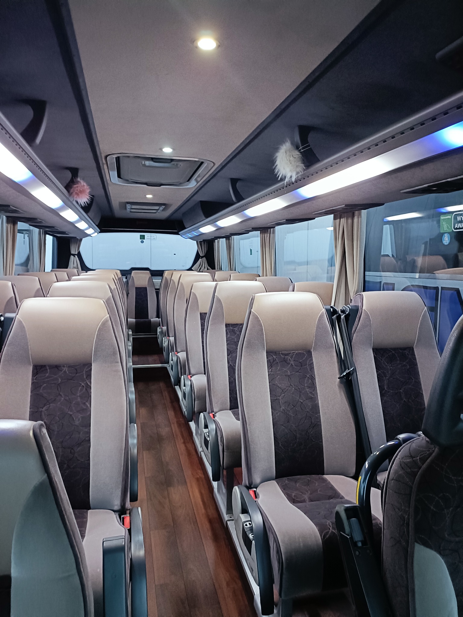 Wnętrze autobusu z rzędami foteli w odcieniach szarości i brązu. Drewnopodobna podłoga, oświetlenie LED i zasłony w oknach. Widok z perspektywy pasażera.