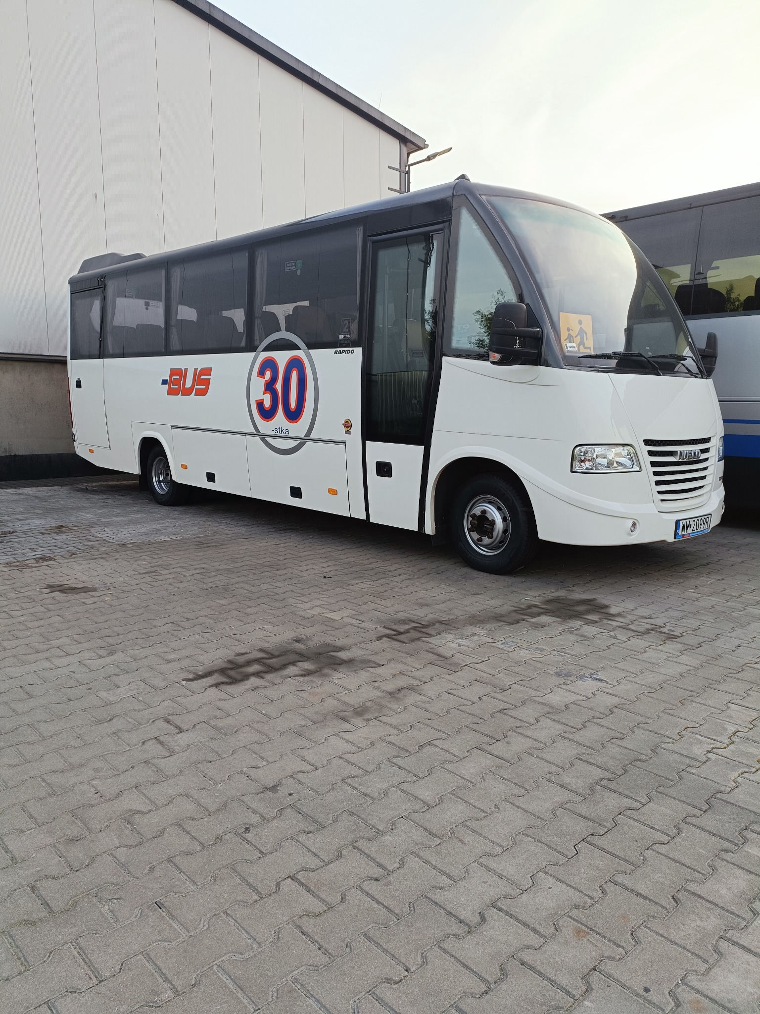 Biały bus z czarnym dachem, oznaczony numerem 30 i napisem BUS, zaparkowany na brukowanym placu przed jasnym budynkiem w słoneczny dzień. Widoczny fragment innego busa.