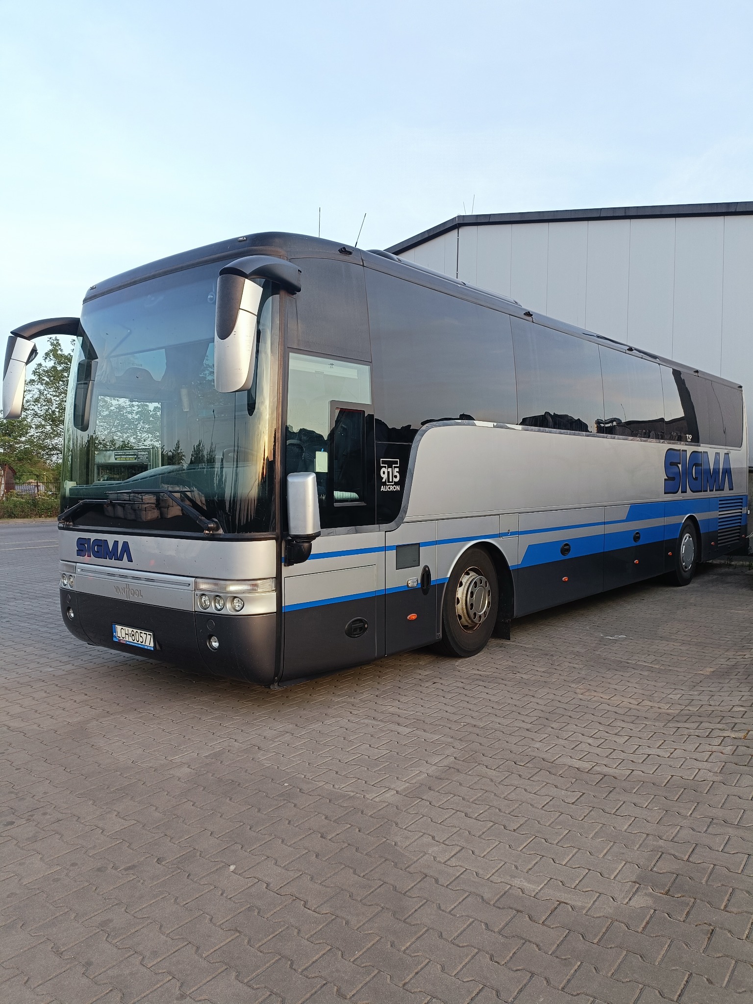 Srebrno-czarny autokar SIGMA z niebieskimi akcentami, zaparkowany na brukowanym placu, widok z boku, tablica rejestracyjna LCH-80577, budynek w tle.