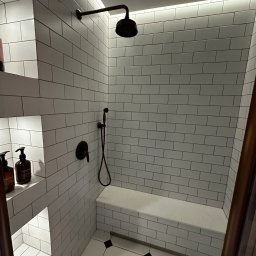 DOKŁADNI.PL SPÓŁKA Z OGRANICZONĄ ODPOWIEDZIALNOŚCIĄ - Nowoczesna kabina prysznicowa z białymi, prostokątnymi płytkami, wnękami na kosmetyki i siedziskiem. Ciemna armatura i oświetlenie LED podkreślają minimalistyczny design.