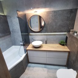 DOKŁADNI.PL SPÓŁKA Z OGRANICZONĄ ODPOWIEDZIALNOŚCIĄ - Nowoczesna łazienka z szarymi płytkami, drewnianym blatem i okrągłym lustrem. Minimalistyczny design z roślinami doniczkowymi jako akcent dekoracyjny. Kabina prysznicowa ze szklaną ścianką.
