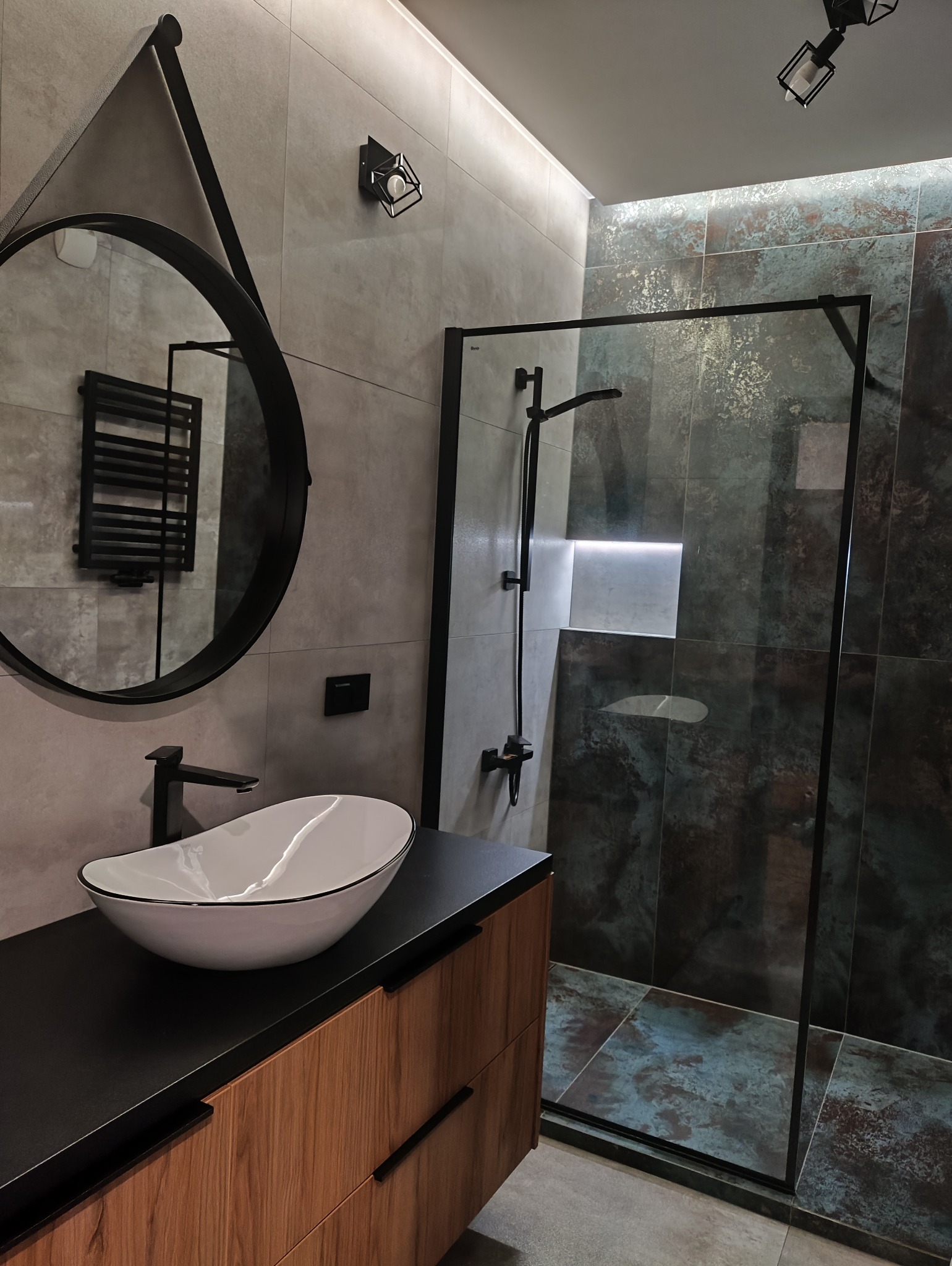 Nowoczesna łazienka z okrągłym lustrem, umywalką nablatową i kabiną prysznicową z ciemnymi płytkami imitującymi rdzę. Minimalistyczne oświetlenie.
