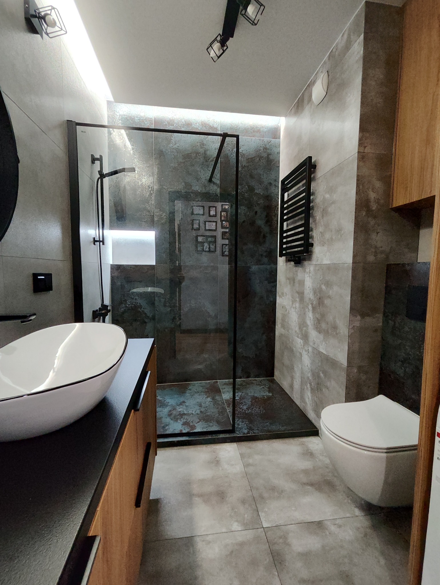 Nowoczesna łazienka z kabiną typu walk-in, umywalką nablatową i grzejnikiem drabinkowym. Płytki imitujące beton w odcieniach szarości i brązu. Minimalistyczne oświetlenie.