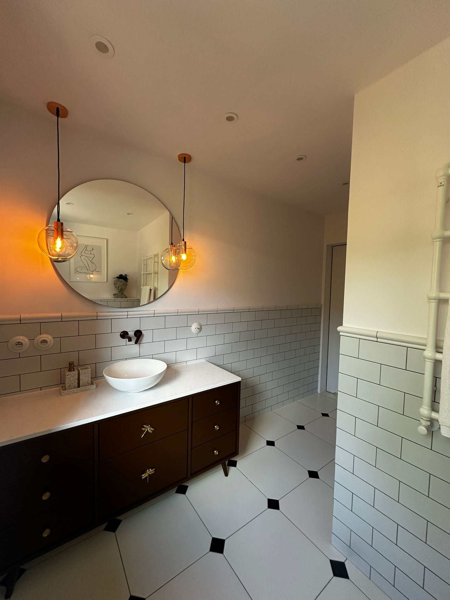 Stylowa łazienka z okrągłym lustrem, wiszącymi lampami i ciemną szafką pod umywalką z ozdobnymi uchwytami w kształcie ważek. Biało-czarna podłoga i ściany z białych płytek.