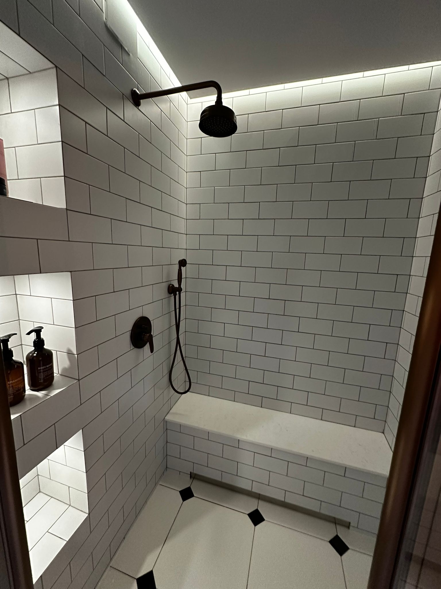 Nowoczesna kabina prysznicowa z białymi, prostokątnymi płytkami, wnękami na kosmetyki i siedziskiem. Ciemna armatura i oświetlenie LED podkreślają minimalistyczny design.