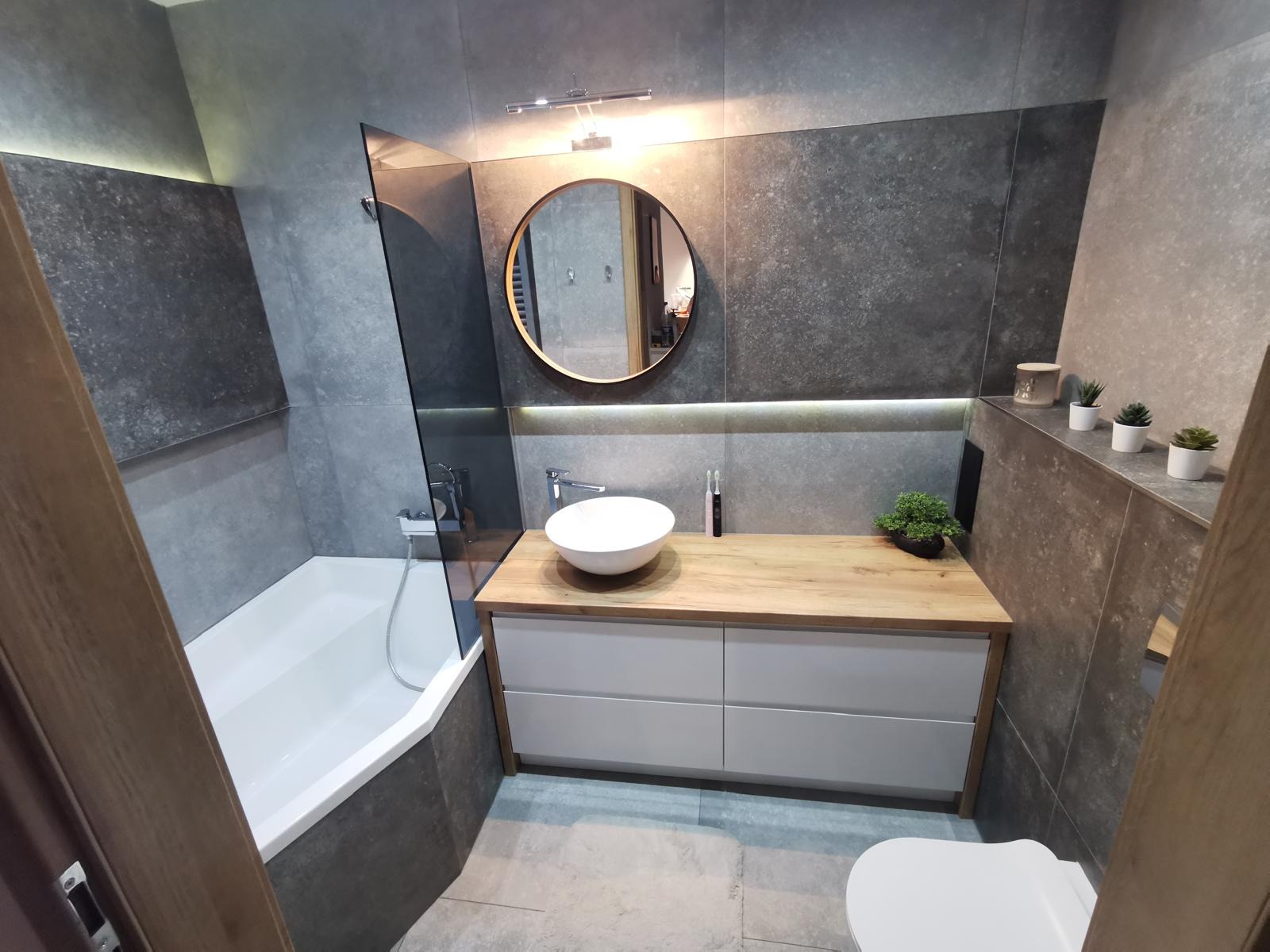 Nowoczesna łazienka z szarymi płytkami, drewnianym blatem i okrągłym lustrem. Minimalistyczny design z roślinami doniczkowymi jako akcent dekoracyjny. Kabina prysznicowa ze szklaną ścianką.