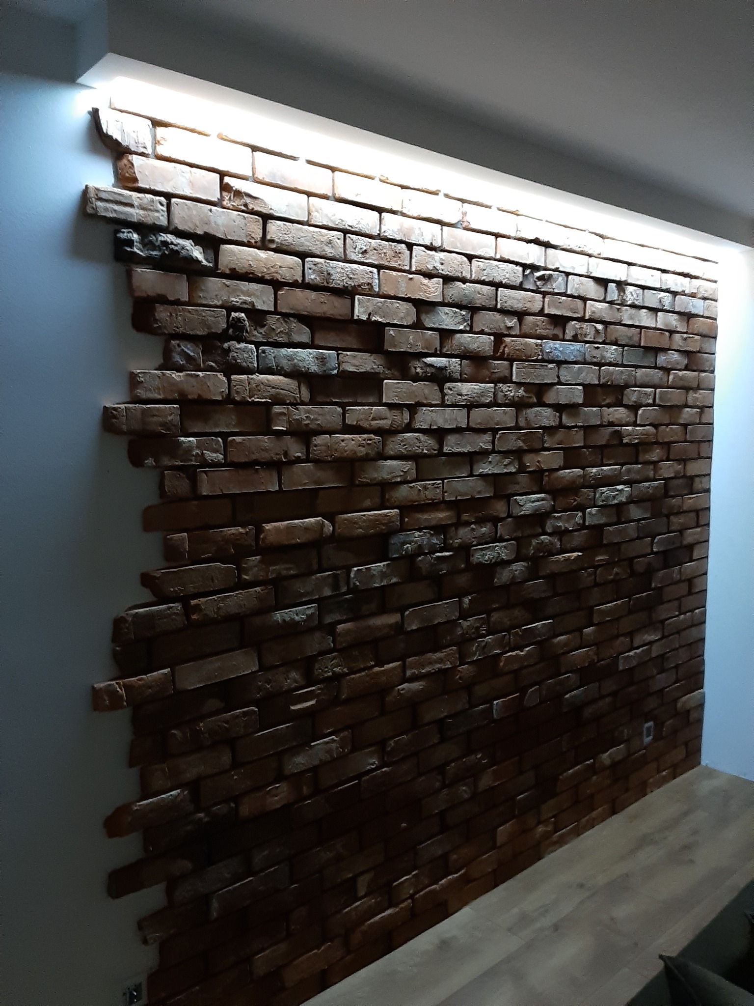 Ściana z cegły dekoracyjnej w salonie, z podświetleniem LED u góry. Widoczna faktura cegieł i fragment drewnianej podłogi. Wyjątkowy efekt dekoracyjny.