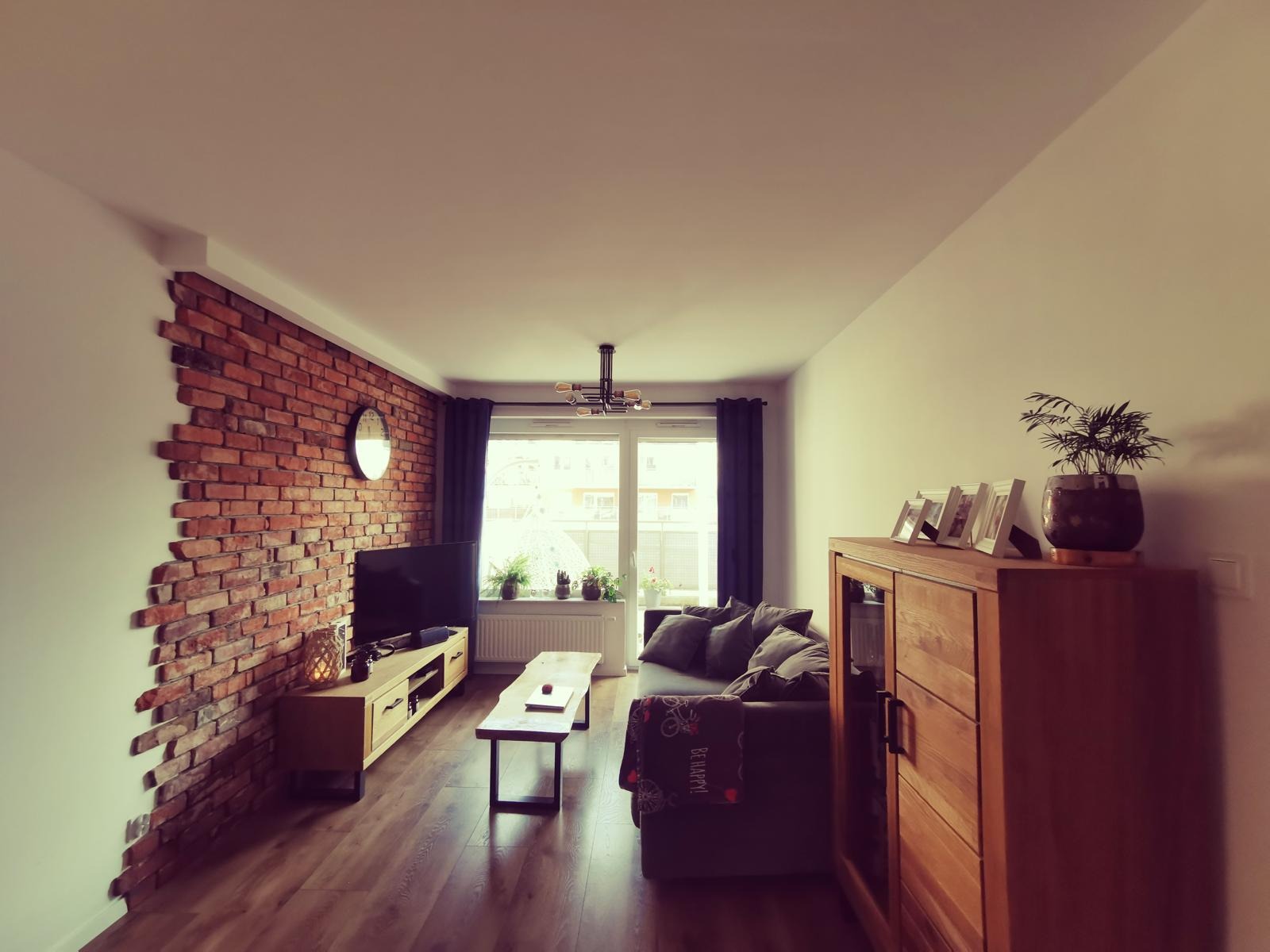 Salon z ceglaną ścianą, drewnianą podłogą i meblami w ciepłych odcieniach. Widoczne okno z zasłonami, kanapa z poduszkami i dekoracje roślinne. Przytulne wnętrze.