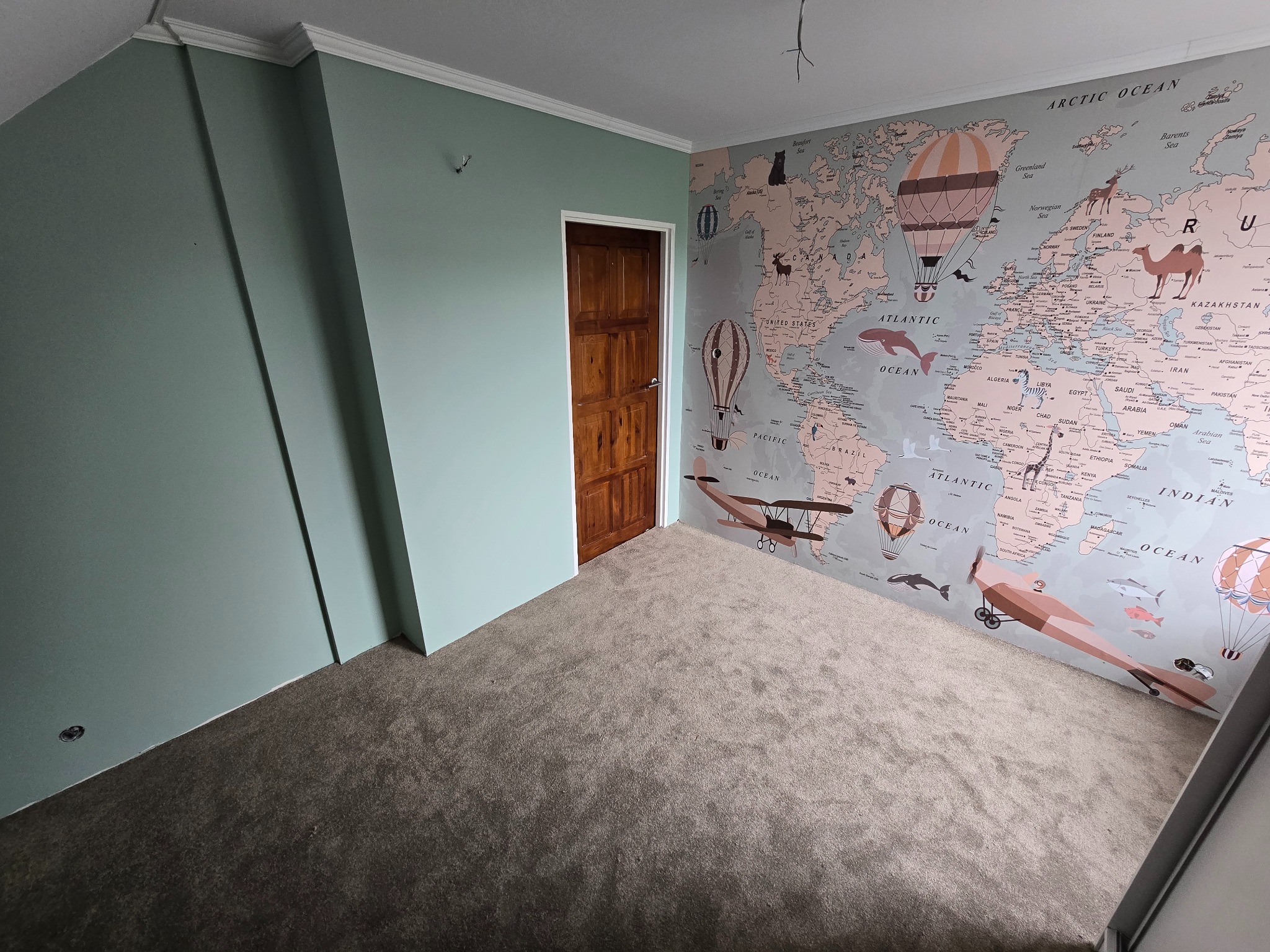 Pokój po remoncie: ściana pomalowana na zielono, drzwi drewniane, tapeta z mapą świata z balonami i samolotami. Nowa, szara wykładzina podłogowa. Biały sufit z listwami.