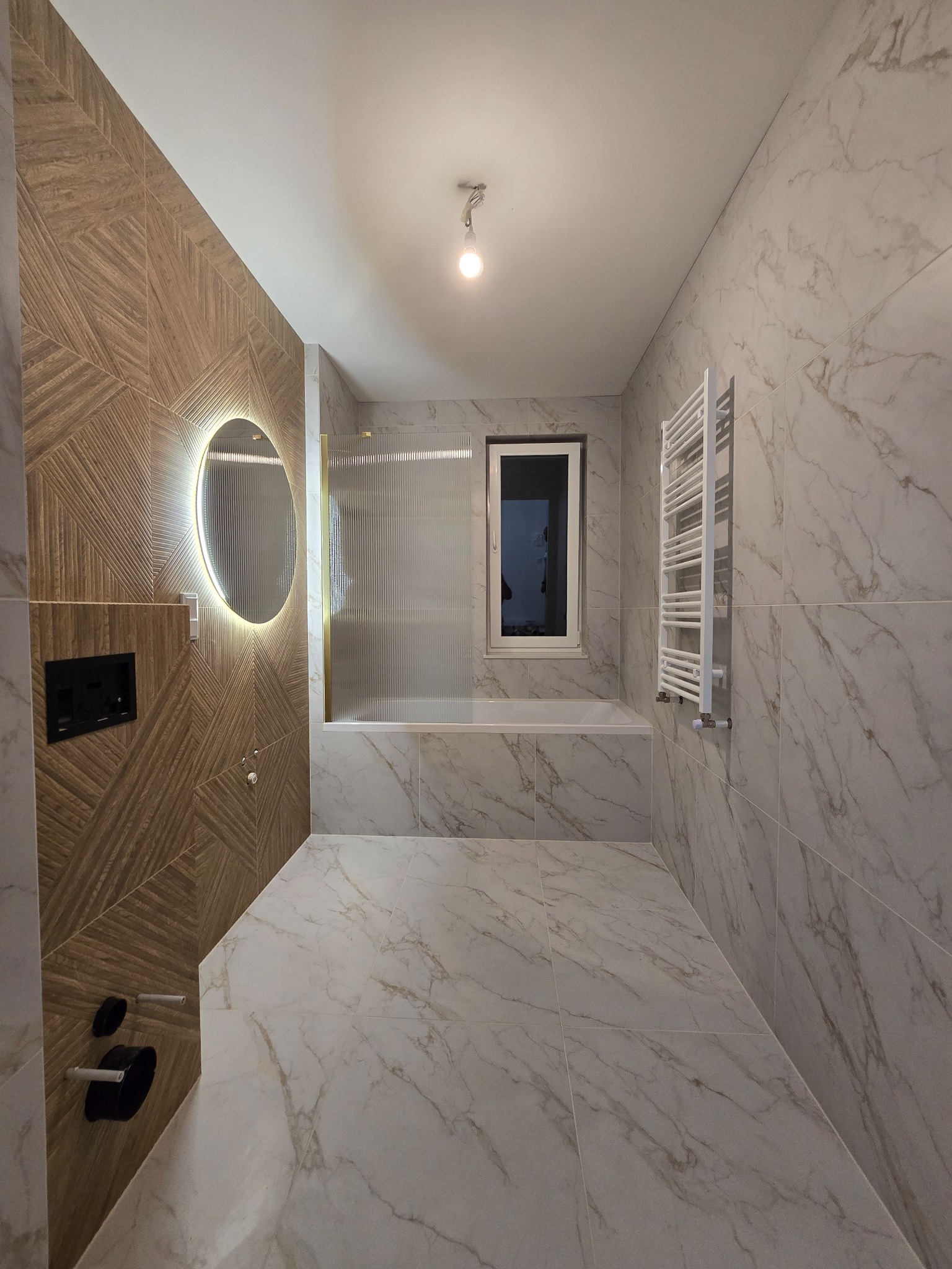 Nowoczesna łazienka z marmurowymi płytkami, oświetlonym lustrem i drewnopodobną ścianą. Widoczna wanna za szklaną ścianką i grzejnik drabinkowy.