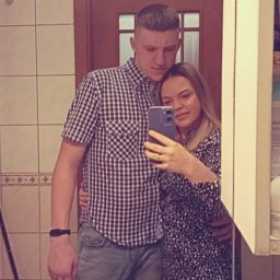 PRZEMYSŁAW ROSIŃSKI - Para robi selfie w łazience z beżowymi płytkami. Mężczyzna w koszuli w kratę, kobieta w sukience w kwiaty. Widoczne umywalka i drzwi.