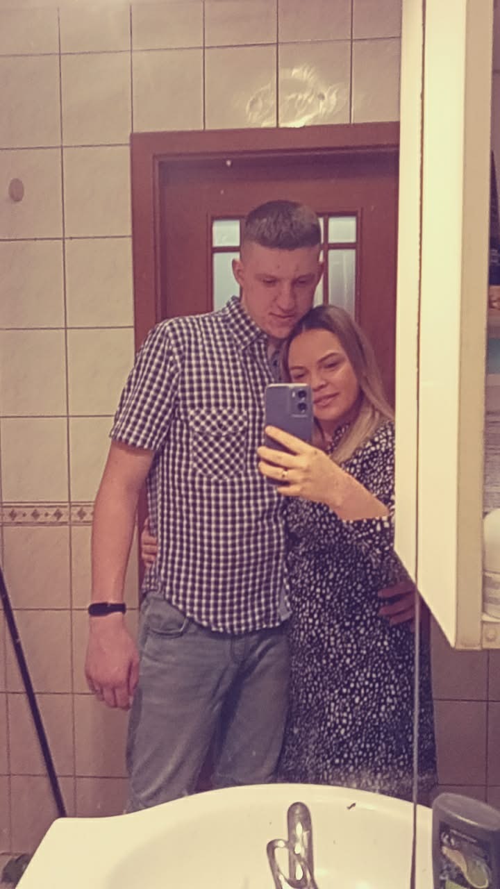 Para robi selfie w łazience z beżowymi płytkami. Mężczyzna w koszuli w kratę, kobieta w sukience w kwiaty. Widoczne umywalka i drzwi.