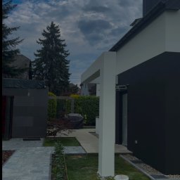 Usługi ogólnobudowlane Wojciech Łukosz ARES-BUD - Nowoczesny dom z antracytową elewacją i jasnymi elementami architektonicznymi. Widoczny fragment ogrodu z kostką brukową i świeżo posadzoną zielenią.