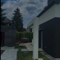 Nowoczesny dom z antracytową elewacją i jasnymi elementami architektonicznymi. Widoczny fragment ogrodu z kostką brukową i świeżo posadzoną zielenią.