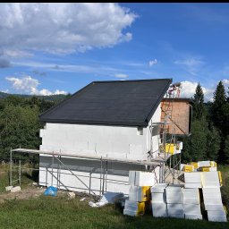 Usługi ogólnobudowlane Wojciech Łukosz ARES-BUD - Budowa domu jednorodzinnego w trakcie ocieplania, widoczne rusztowania, białe bloczki, ocieplenie i pracownik na dachu na tle zieleni i błękitnego nieba.