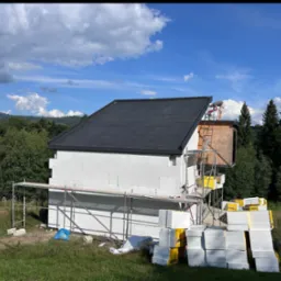 Budowa domu jednorodzinnego w trakcie ocieplania, widoczne rusztowania, białe bloczki, ocieplenie i pracownik na dachu na tle zieleni i błękitnego nieba.