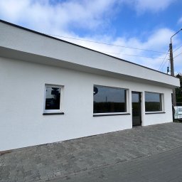 Usługi ogólnobudowlane Wojciech Łukosz ARES-BUD - Biały budynek z oknami i czarnym dachem na tle błękitnego nieba. Widoczny chodnik z kostki brukowej i fragment ulicy. Minimalistyczna architektura budynku.